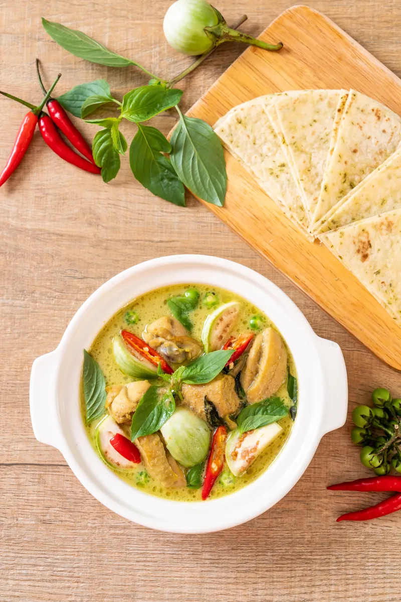 Sumptuous green curry in a white bowl, garnished with fresh Thai basil and vibrant red chili slices, set against a rustic wooden background.